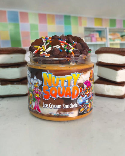 A jar of Nutty Squad Ice Cream Sandwich spread sits on a light countertop, flanked by two stacked ice cream sandwich halves. The jar has a colorful cartoon label and is topped with chocolate pieces and rainbow sprinkles.