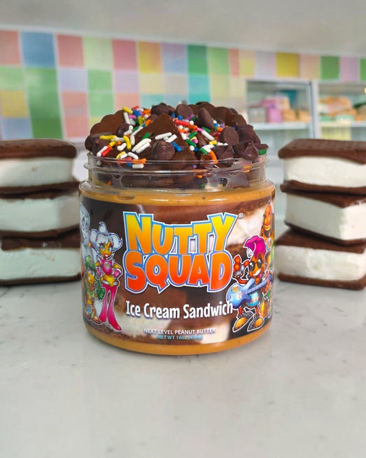 A jar of Nutty Squad Ice Cream Sandwich spread sits on a light countertop, flanked by two stacked ice cream sandwich halves. The jar has a colorful cartoon label and is topped with chocolate pieces and rainbow sprinkles.