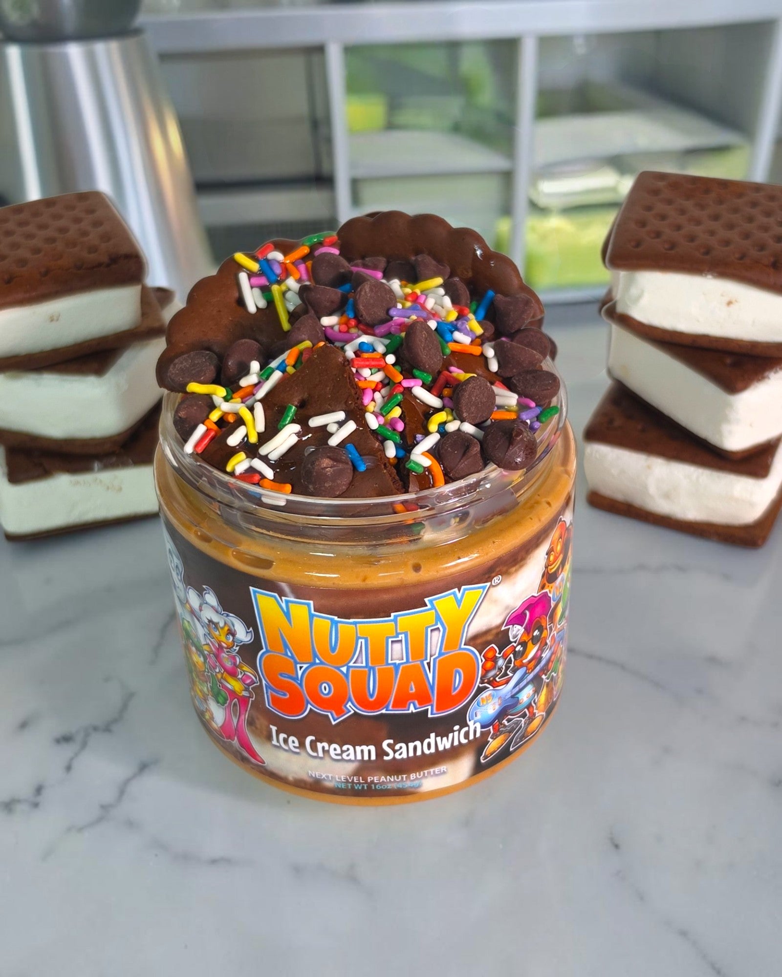 A jar of Nutty Squad Ice Cream Sandwich filled with chocolate ice cream and topped with chocolate chips and rainbow sprinkles, displayed on a marble countertop with stacks of the sandwich on both sides.