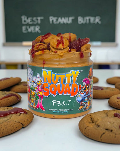 A jar of Nutty Squad PB & J peanut butter sits on a white table surrounded by cookies, with a chalkboard in the background reading 'BEST PEANUT BUTTER EVER'.